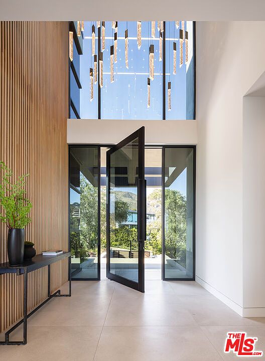 This grand, modern entryway features a dramatic floor-to-ceiling glass pivot door that floods the space with natural light. The interior is characterized by a striking wood-slat accent wall, a minimalist console table with a decorative plant, and a sophisticated, multi-tiered crystal chandelier hanging from the high ceiling. The clean lines, neutral tile flooring, and seamless transition to the outdoor landscape create an atmosphere of luxury and architectural elegance.