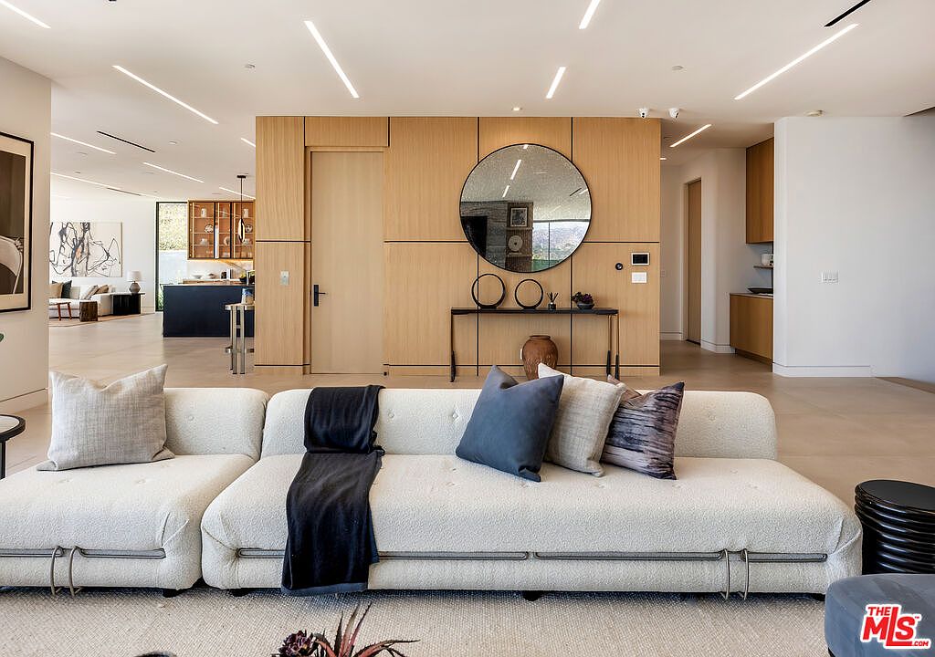 This sophisticated living room features a large, textured cream-colored sectional sofa adorned with plush throw pillows and a dark throw blanket. The space is anchored by a minimalist wood-paneled wall featuring a circular mirror and a sleek console table, creating a clean, contemporary aesthetic. The open-concept design provides a clear view into the adjacent kitchen, emphasizing the home's spacious and airy layout.