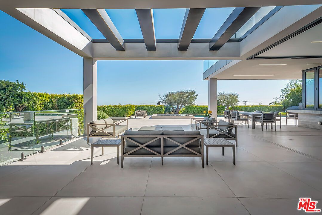 This expansive, modern covered patio offers a seamless transition between indoor and outdoor living, featuring sleek gray tile flooring and a minimalist pergola structure. The space is furnished with comfortable, contemporary outdoor seating and a dining area, all overlooking a pristine pool and lush, manicured landscaping. The perspective captures the open-air design, emphasizing the bright, airy atmosphere and the luxurious, resort-like feel of the property.