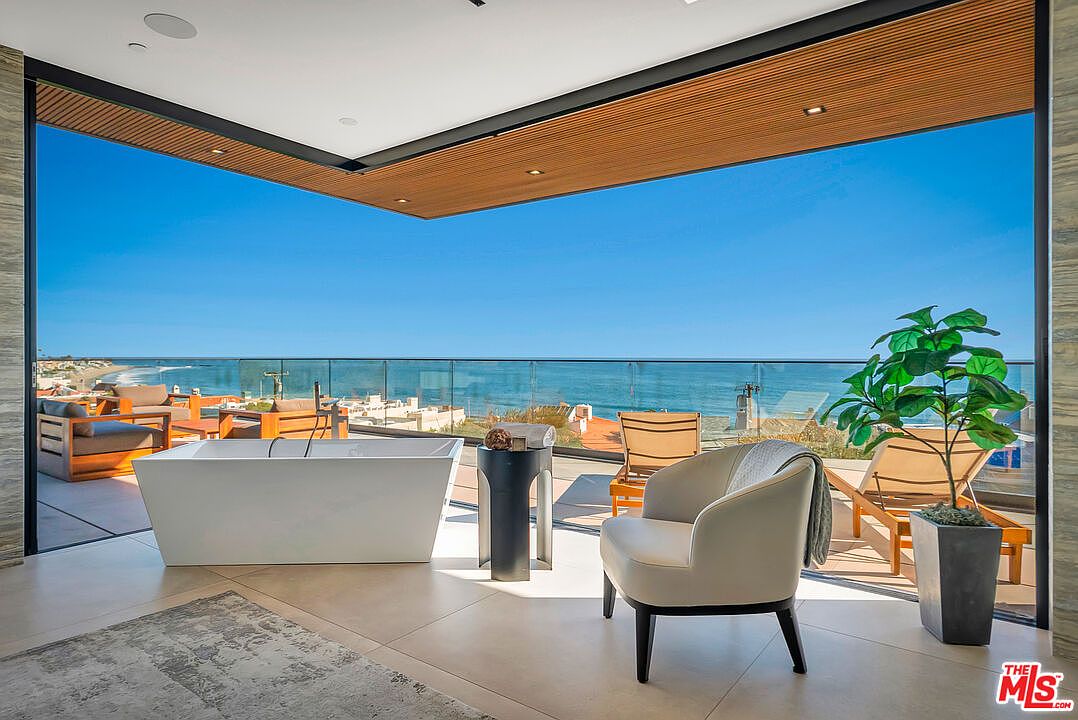 This luxurious primary bathroom features an open-concept design with a sleek, freestanding white soaking tub positioned to overlook a breathtaking panoramic ocean view. The space seamlessly transitions to an expansive outdoor balcony through floor-to-ceiling glass walls, creating an indoor-outdoor living experience. A modern armchair and minimalist side table complete the serene, high-end aesthetic of this coastal retreat.