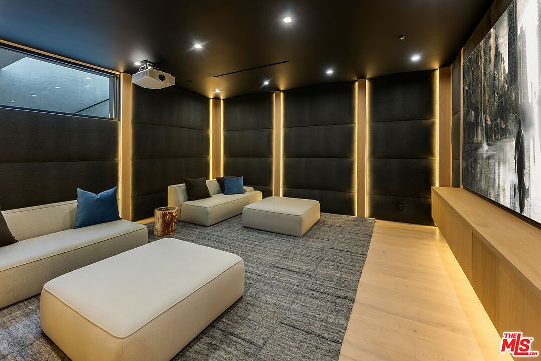 This sophisticated home theater features plush, low-profile modular seating in a neutral palette, accented by vibrant blue throw pillows and a rustic wooden side table. The room is defined by dark, upholstered wall panels with integrated vertical LED lighting, creating a moody and cinematic atmosphere. A large projection screen dominates the front wall, complemented by a sleek, light-wood media console and a projector mounted on the dark ceiling.