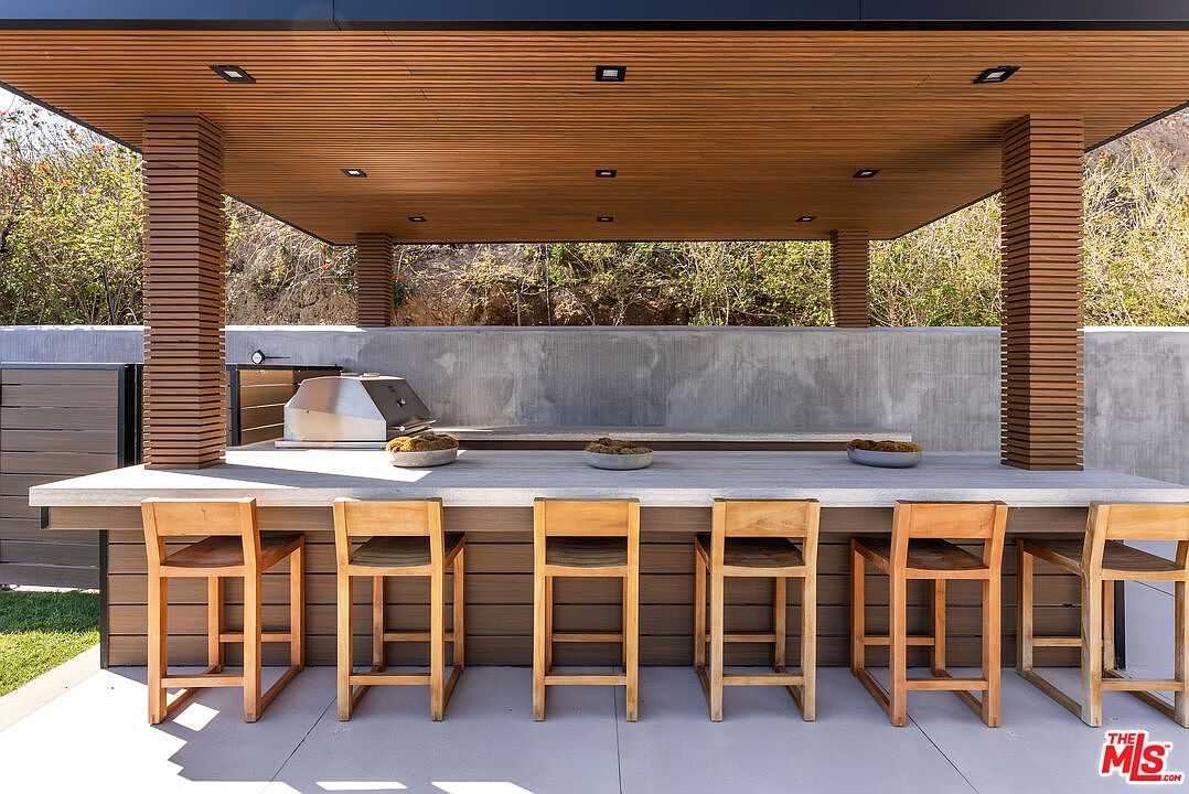 This sophisticated outdoor kitchen and bar area features a sleek concrete countertop supported by a warm wood-slatted base and matching bar stools. The space is sheltered by a modern, slatted wood ceiling with recessed lighting, set against a clean concrete wall backdrop. The overall aesthetic is contemporary and minimalist, perfect for high-end outdoor entertaining.