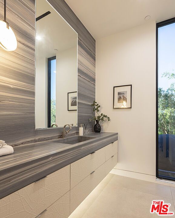 This modern powder room features a striking vanity topped with dramatic, horizontally-veined grey stone that extends up the wall to frame a large, frameless mirror. Below the stone counter, textured white cabinetry adds a subtle geometric pattern, while under-cabinet lighting creates a floating effect. The space is brightened by a tall, floor-to-ceiling window that brings in natural light and a view of the outdoors, complemented by a minimalist pendant light.