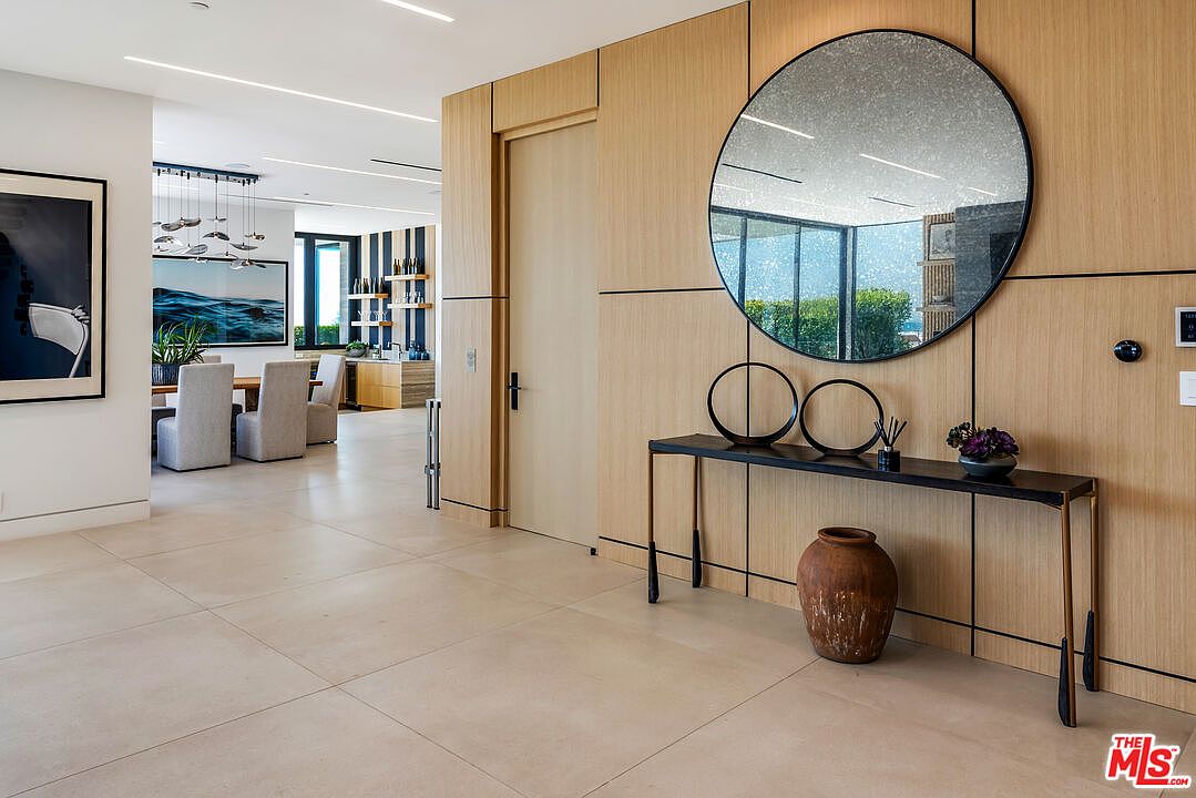 This elegant entryway features light wood paneling, a large circular mirror, and a sleek console table adorned with minimalist decor and a rustic clay vase. The perspective looks from the foyer into a bright, open-concept dining area, showcasing a sophisticated modern aesthetic with clean lines and neutral tones. The space feels airy and luxurious, emphasizing high-end finishes and a seamless transition between living areas.
