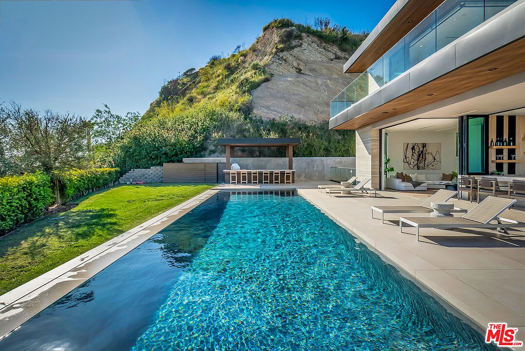 This stunning modern luxury estate features a sleek, rectangular infinity-edge pool as the centerpiece of its expansive outdoor living area. The scene includes a covered outdoor bar with seating, a manicured lawn, and contemporary lounge chairs on a spacious patio, all set against a dramatic, rugged hillside backdrop. The seamless transition between the indoor living space and the outdoor terrace highlights a sophisticated, high-end architectural design.