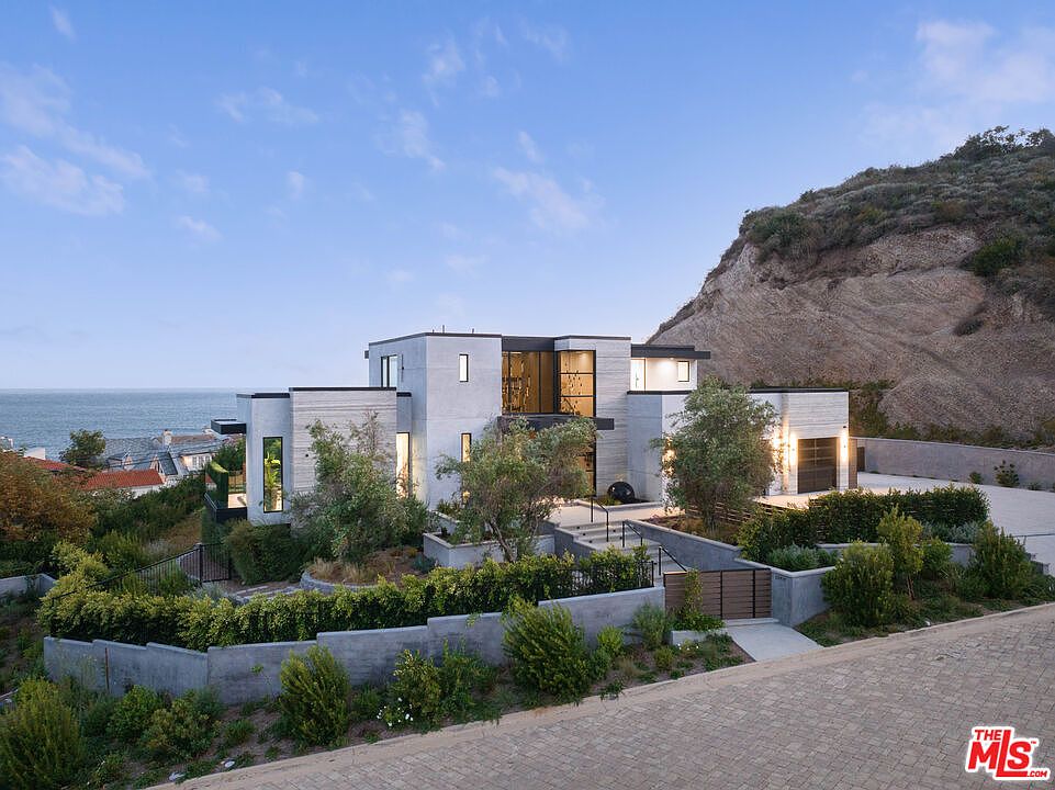 This modern, multi-level luxury residence is nestled against a rugged hillside, showcasing a sophisticated architectural design with clean lines and a neutral stone facade. The property features a tiered landscape with lush greenery, a paved driveway, and a prominent entryway, all set against a stunning coastal backdrop. The perspective captures the home's integration with its natural environment, emphasizing its exclusive and serene location.