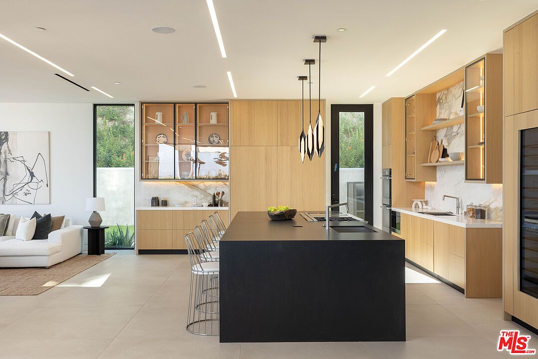 This modern, open-concept kitchen features a striking matte black central island contrasted by warm, light-toned wood cabinetry and marble backsplashes. The space is illuminated by sleek, recessed linear lighting and elegant pendant fixtures, creating a sophisticated and airy atmosphere. The perspective captures the seamless flow between the culinary workspace and the adjacent living area, highlighting the high-end finishes and contemporary design.