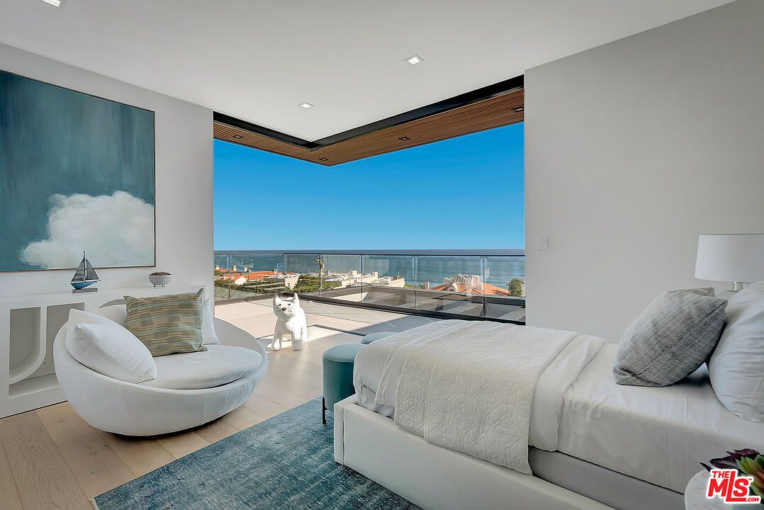 This luxurious primary bedroom features a contemporary design with a seamless transition to a private balcony through expansive floor-to-ceiling glass walls. The room is anchored by a plush white bed and a stylish circular lounge chair, all set against a backdrop of stunning ocean views. The minimalist aesthetic, combined with high-end finishes and a serene color palette, creates an atmosphere of sophisticated coastal elegance.