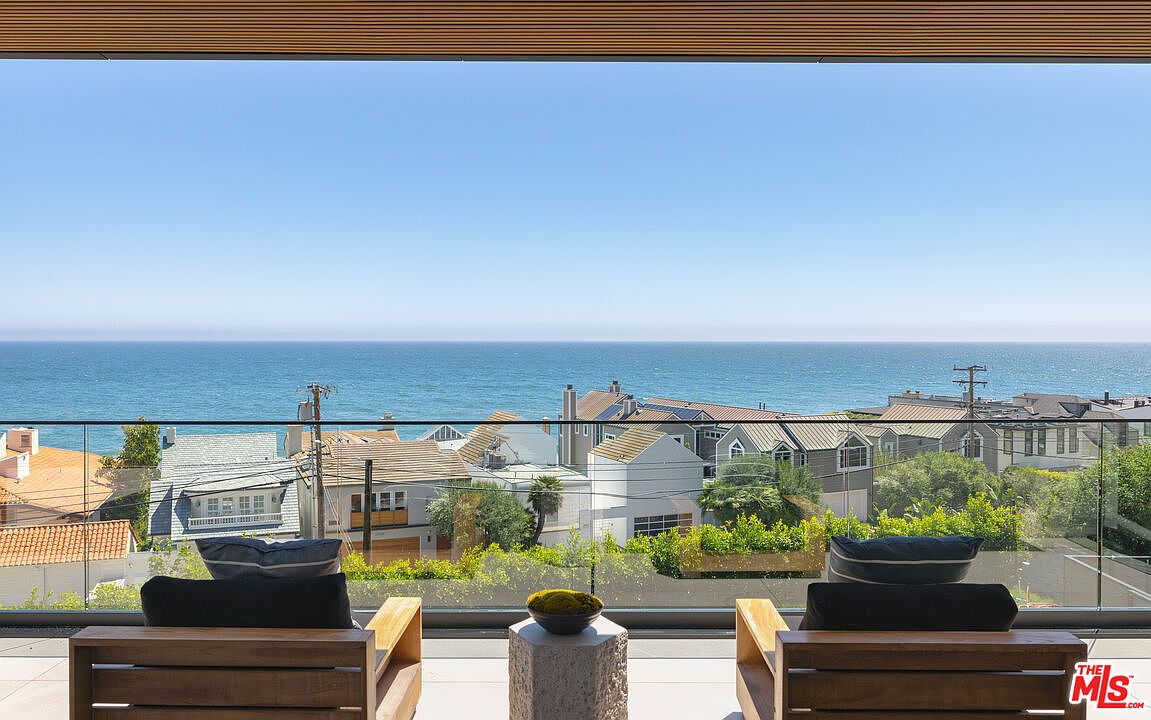 This image captures a stunning, modern balcony overlooking the Pacific Ocean, featuring two sleek wooden lounge chairs with dark cushions and a central stone side table. The minimalist glass railing provides an unobstructed, panoramic view of the coastal neighborhood and the vast horizon beyond. The scene evokes a sense of luxury, tranquility, and high-end coastal living.