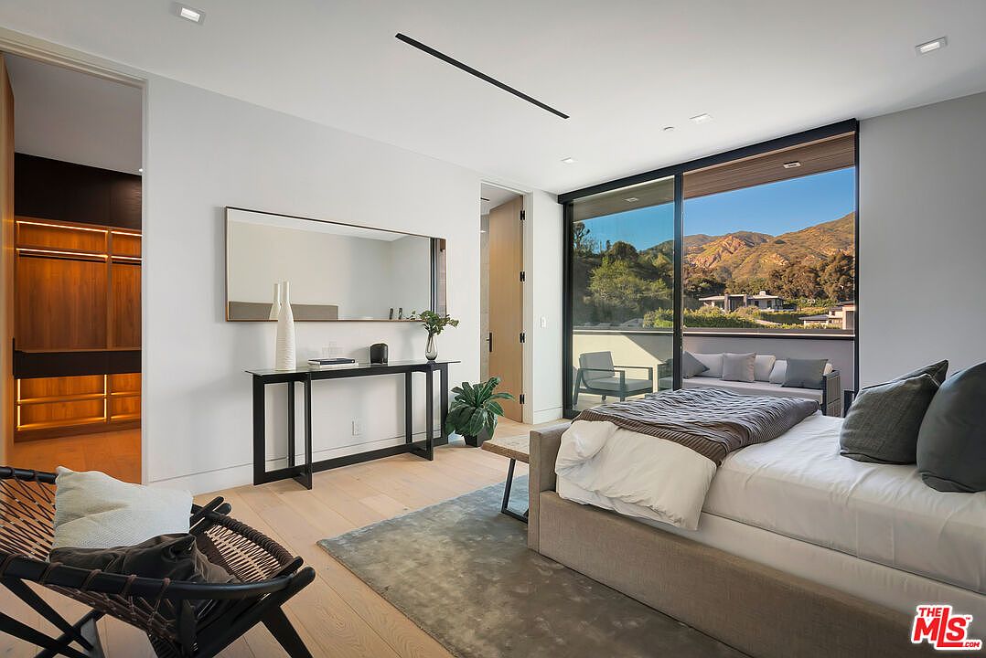 This luxurious primary bedroom features a contemporary design with light hardwood floors, a neutral color palette, and a large sliding glass door that opens to a private balcony with scenic mountain views. The room is furnished with a plush bed, a sleek console table beneath a large wall mirror, and a walk-in closet visible in the background. The space feels airy and sophisticated, emphasizing a seamless connection between indoor comfort and the natural landscape.