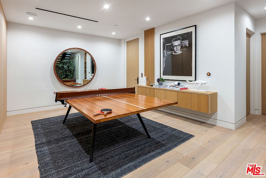 This modern game room features a sleek, wooden ping pong table as the centerpiece, set atop a dark textured area rug. The space is accented by a large circular mirror, a floating wooden console table adorned with books and a decorative shark sculpture, and a prominent framed portrait on the wall. The room is illuminated by recessed lighting and maintains a clean, minimalist aesthetic with light hardwood flooring.