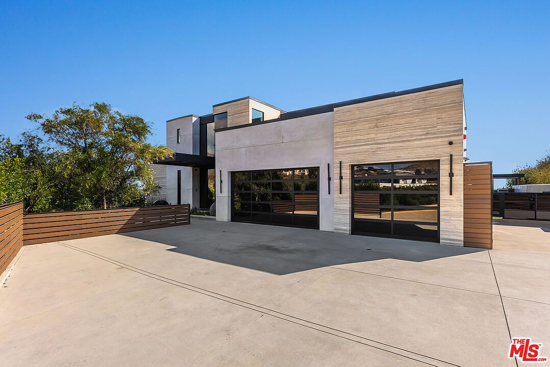 This modern architectural home features a prominent two-car garage with sleek, black-framed glass panel doors. The exterior showcases a sophisticated blend of smooth concrete and horizontal stone cladding, set against a clear blue sky. A wide, clean concrete driveway leads up to the structure, framed by contemporary wooden fencing and mature landscaping.