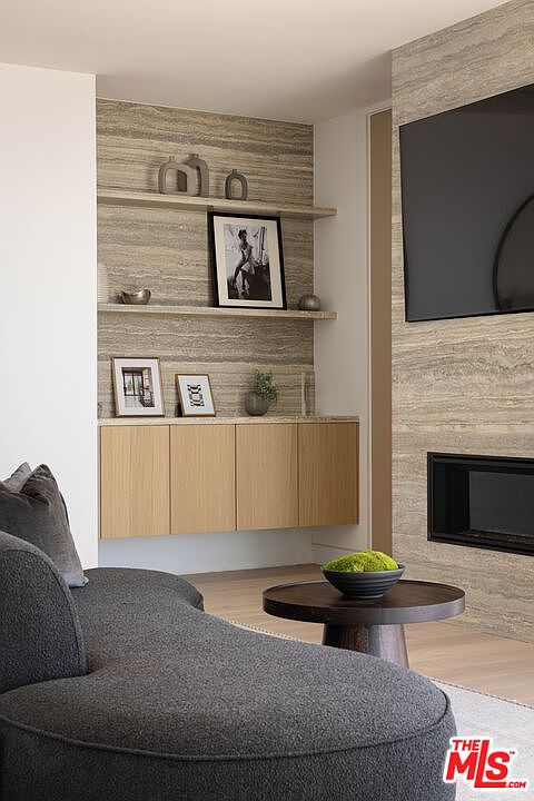 This sophisticated living room features a textured, stone-clad accent wall that integrates a fireplace and custom built-in shelving. A plush, charcoal-colored curved sofa anchors the space, complemented by a minimalist dark wood coffee table topped with a decorative bowl of greenery. The room exudes a modern, serene aesthetic with its neutral color palette and clean, architectural lines.