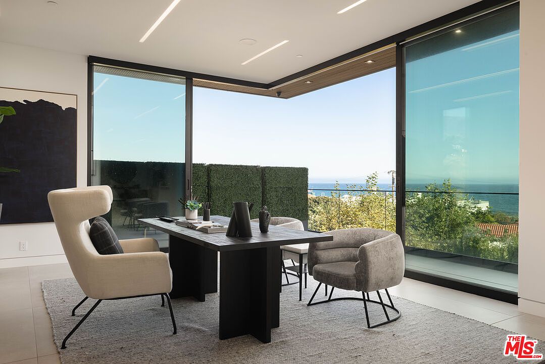 This sophisticated home office features a sleek, dark wood desk positioned to take full advantage of the panoramic ocean views through expansive floor-to-ceiling glass walls. The room is furnished with a contemporary cream-colored wingback chair and a plush grey velvet guest chair, all set atop a textured area rug. The minimalist design, combined with recessed linear lighting and a seamless indoor-outdoor transition, creates an atmosphere of professional luxury and tranquility.