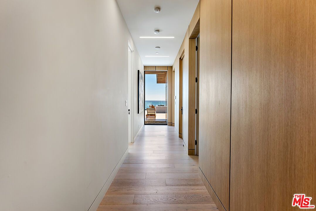 This sleek, minimalist hallway features light-toned wood flooring and matching wood-paneled walls on the right, creating a warm and cohesive aesthetic. The perspective draws the eye down the corridor toward a bright, open doorway that reveals a stunning ocean view and a sunlit patio. Recessed ceiling lighting provides a clean, modern look, emphasizing the architectural simplicity and high-end finish of the space.