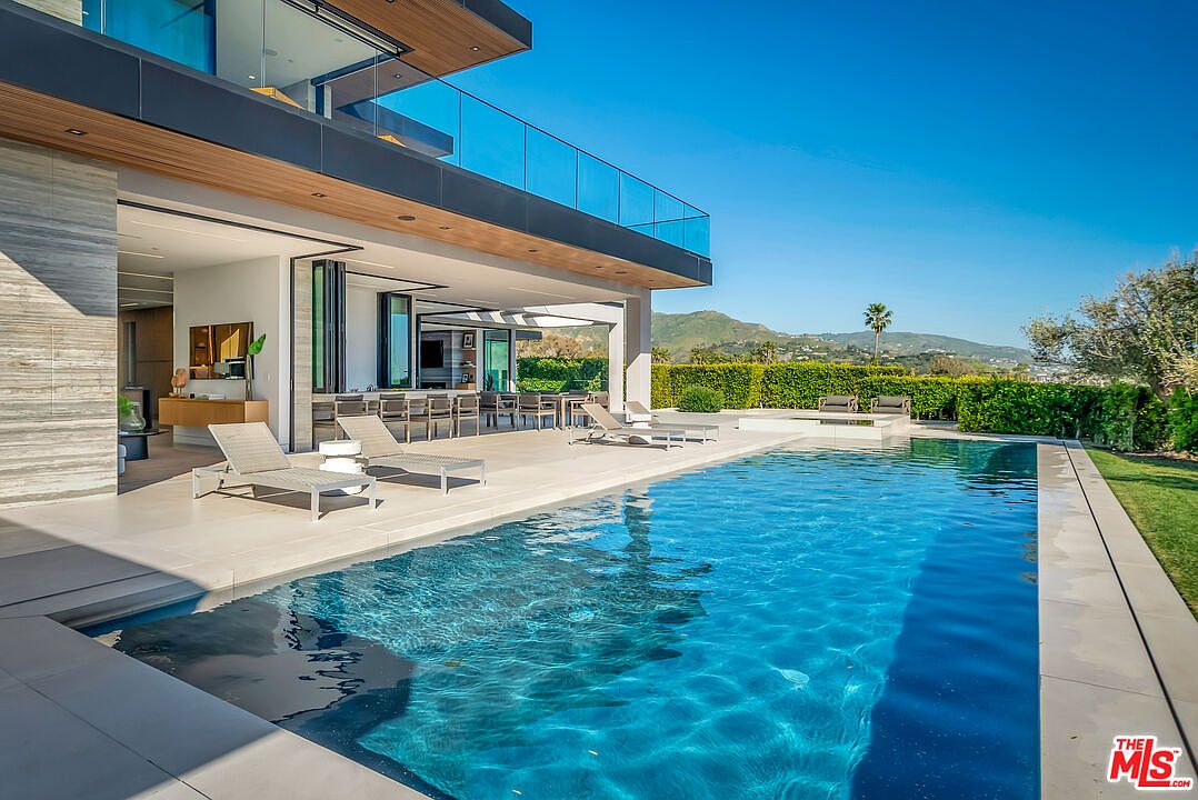 This luxurious modern residence features a seamless indoor-outdoor flow, highlighted by a stunning infinity-edge swimming pool that reflects the clear blue sky. The expansive patio area is furnished with sleek lounge chairs and an outdoor dining space, all set against a backdrop of manicured hedges and distant rolling hills. The architectural design emphasizes clean lines, glass railings, and high-end materials, creating an atmosphere of sophisticated relaxation.