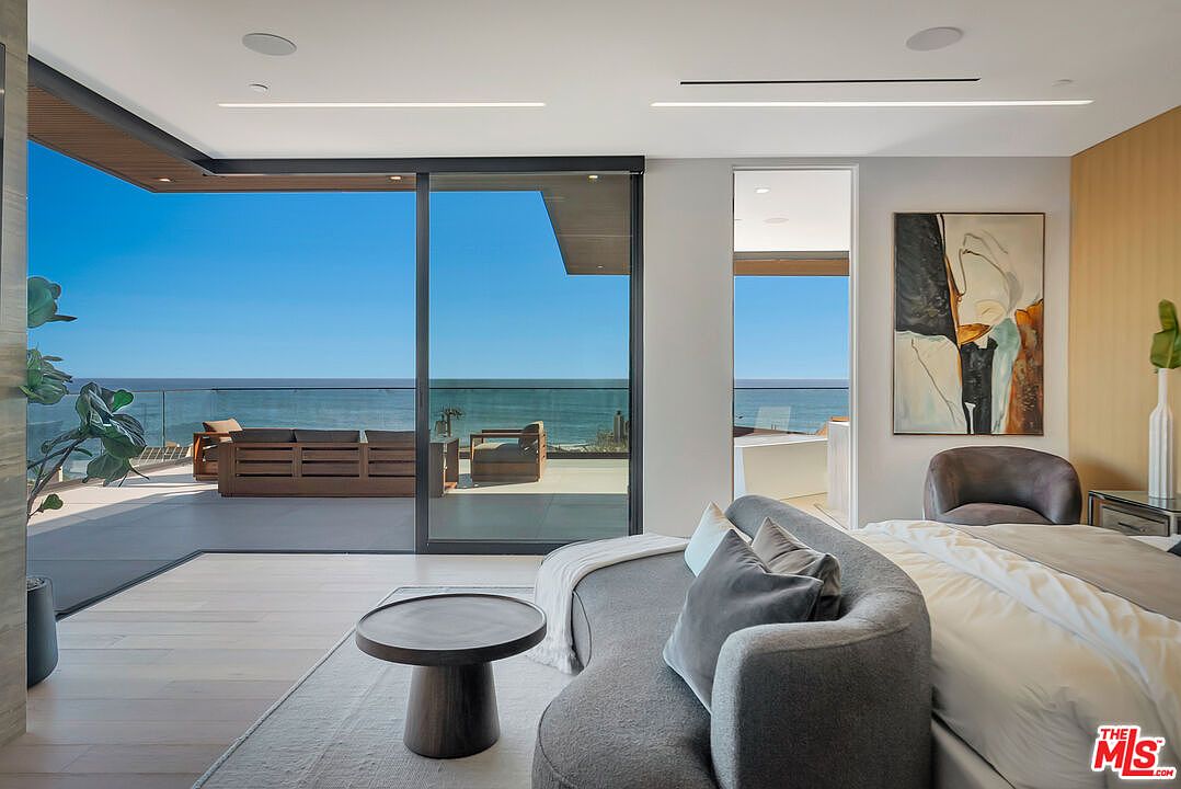 This luxurious primary bedroom features a sophisticated, modern aesthetic with a curved grey sofa positioned at the foot of the bed. Large floor-to-ceiling sliding glass doors open onto a spacious balcony, offering an expansive, unobstructed view of the ocean. The room is accented by a large abstract painting and minimalist decor, creating a serene and high-end coastal retreat.
