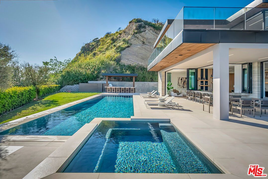 This stunning modern luxury estate features a seamless indoor-outdoor flow, highlighted by a sleek infinity-edge pool and a built-in spa overlooking a manicured lawn and hillside. The expansive patio area is furnished with contemporary lounge chairs and an outdoor dining setup, all sheltered by the home's cantilevered upper level. The architectural design emphasizes clean lines, glass railings, and high-end finishes, creating a sophisticated and serene atmosphere perfect for entertaining.