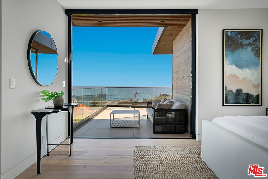 This elegant primary bedroom features a seamless transition to a private balcony through expansive floor-to-ceiling sliding glass doors, offering a stunning panoramic view of the ocean. The interior is decorated with a minimalist aesthetic, including a sleek black console table, a circular mirror, and a large abstract art piece, all set against light hardwood flooring. The overall atmosphere is one of sophisticated coastal luxury, emphasizing natural light and indoor-outdoor living.