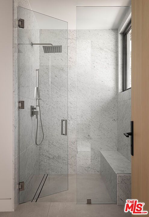 This modern, minimalist walk-in shower features floor-to-ceiling marble tiling and a sleek glass enclosure. The space includes a built-in marble bench, a rainfall showerhead, and a handheld sprayer, creating a luxurious spa-like atmosphere. The perspective is a direct, eye-level shot looking into the shower stall, highlighting the clean lines and high-end materials.