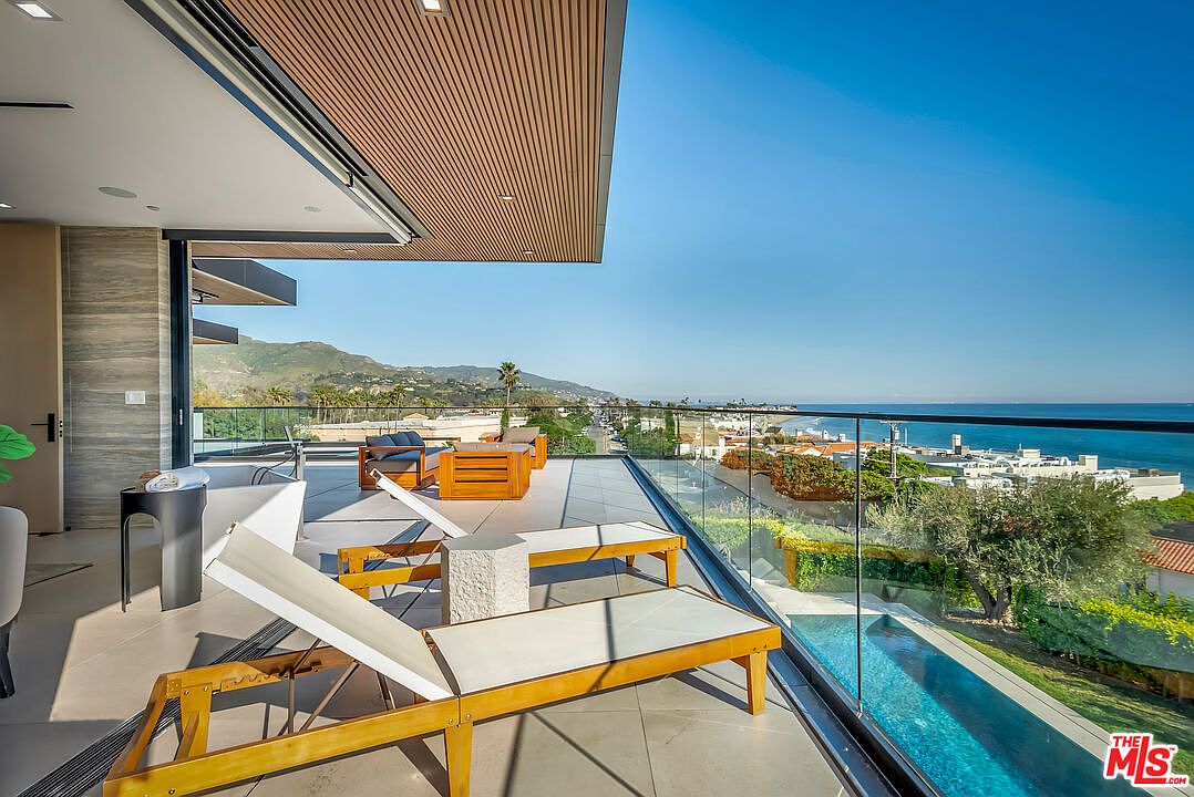 This expansive, modern balcony offers a seamless indoor-outdoor living experience with a stunning panoramic view of the coastline and ocean. The space is furnished with sleek wooden lounge chairs and a comfortable seating area, all protected by a minimalist glass railing that maximizes the horizon view. The architectural design features a warm, slatted wood ceiling and clean lines, creating a luxurious and serene atmosphere perfect for relaxation.
