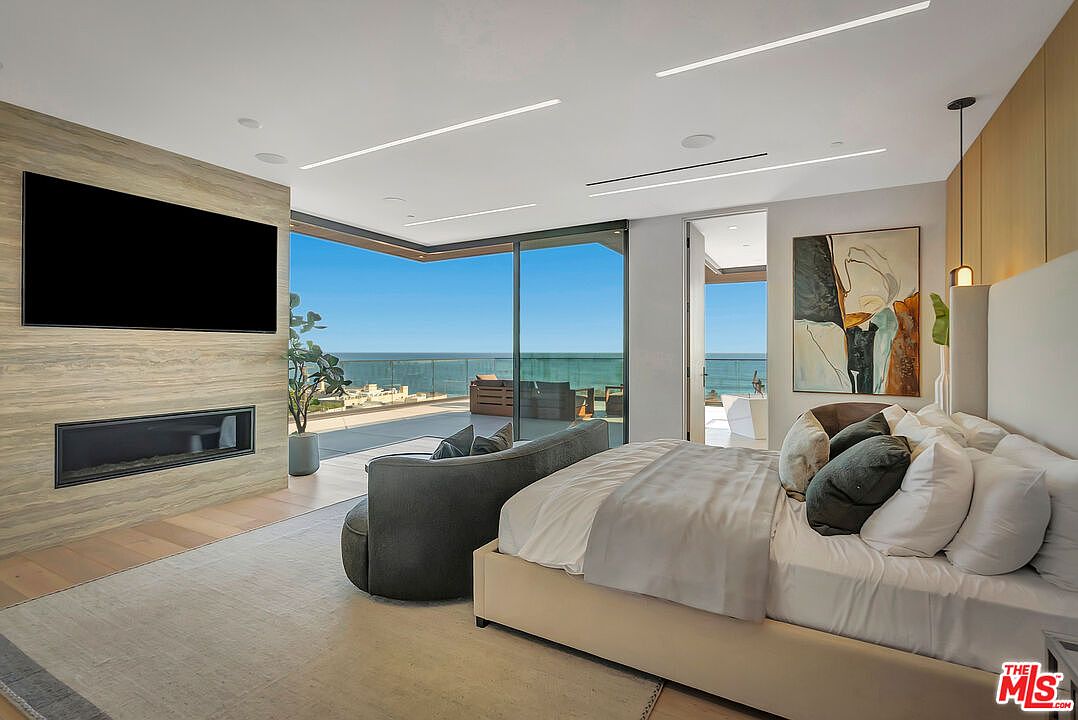 This luxurious primary bedroom features a contemporary design with a neutral color palette, highlighted by a stone-clad fireplace wall and a large flat-screen television. The room seamlessly transitions to a spacious balcony through floor-to-ceiling glass sliding doors, offering an expansive, unobstructed view of the ocean. A plush, low-profile bed and a curved sofa create a sophisticated and relaxing atmosphere, perfectly capturing the essence of high-end coastal living.