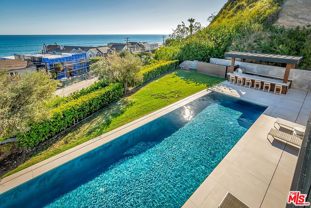 This elevated perspective captures a luxurious backyard oasis featuring a sleek, rectangular infinity-edge swimming pool as the centerpiece. The space includes a manicured lawn, a covered outdoor kitchen with bar seating, and a paved lounge area, all set against a backdrop of coastal cliffs and the Pacific Ocean. The scene exudes a high-end, serene atmosphere perfect for outdoor entertaining and relaxation.