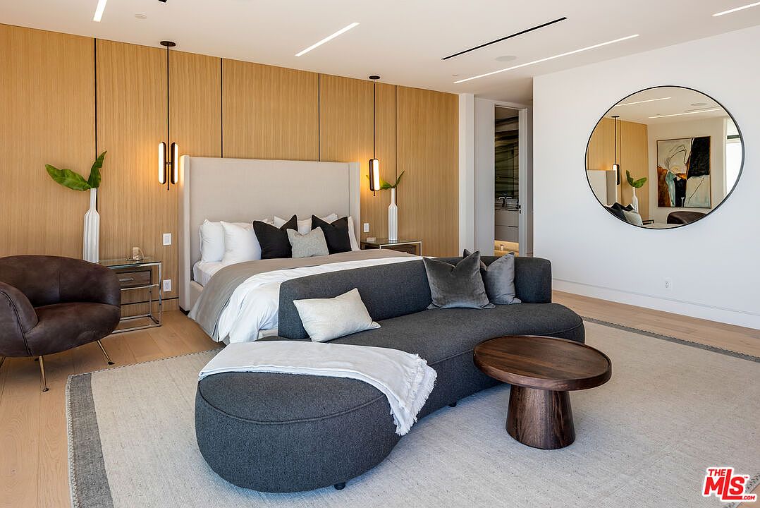 This luxurious primary bedroom features a sophisticated contemporary design with a prominent wood-paneled accent wall behind a plush, light-colored upholstered bed. A curved, dark charcoal sofa sits at the foot of the bed, complemented by a round wooden coffee table and a large circular mirror that reflects the elegant space. The room is bathed in soft, warm lighting from modern pendant fixtures, creating an inviting and high-end atmosphere.