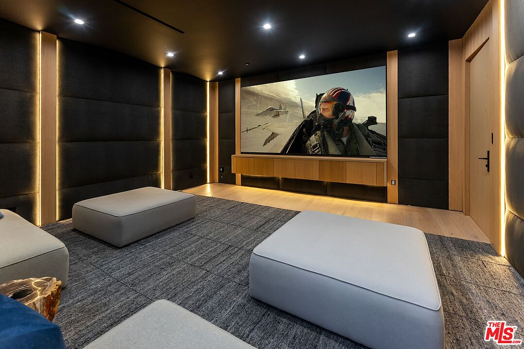 This image showcases a sophisticated, modern home theater designed for an immersive cinematic experience. The room features dark, upholstered wall panels accented by vertical LED strip lighting, a large projection screen displaying a high-action film, and minimalist, low-profile seating on a textured gray carpet. The combination of warm wood cabinetry and sleek, dark finishes creates a luxurious and cozy atmosphere perfect for entertainment.