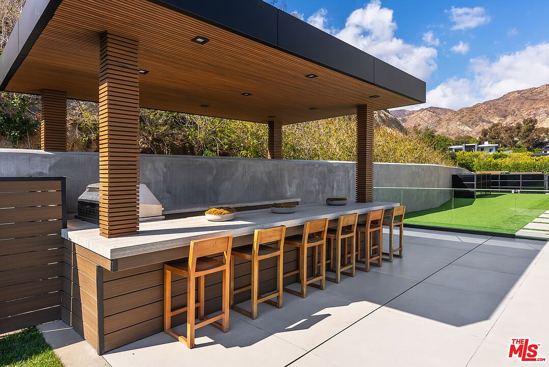 This modern outdoor kitchen and bar area features a sleek concrete countertop, a built-in grill, and a row of wooden bar stools under a contemporary slatted wood-paneled roof. The space is set against a minimalist concrete wall with a glass railing overlooking a manicured lawn and scenic mountain views. The overall aesthetic is sophisticated, clean, and designed for high-end outdoor entertaining.