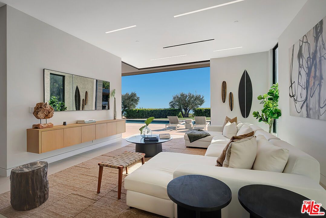 This sophisticated, modern living room features an open-concept design that seamlessly transitions to an outdoor pool area through expansive floor-to-ceiling glass doors. The space is anchored by a large, plush white sectional sofa, a natural fiber area rug, and a sleek, wall-mounted light wood media console. The minimalist aesthetic is complemented by curated decor, including surfboard-inspired wall art and a striking tree-stump side table, creating a serene and luxurious atmosphere.
