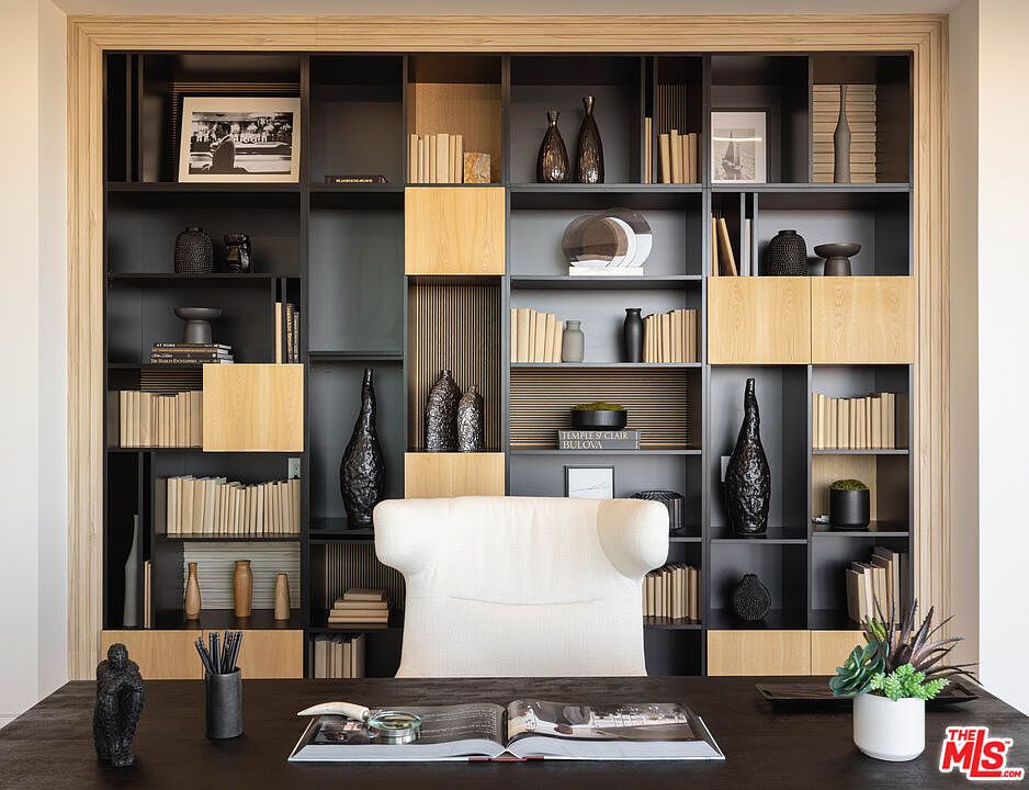 This sophisticated home office features a large, dark wood desk centered in front of a floor-to-ceiling custom shelving unit. The shelving combines black matte finishes with warm wood accents, creating a modern and organized aesthetic filled with curated decor, books, and vases. A plush, white upholstered chair provides a comfortable focal point, emphasizing a professional yet inviting workspace.