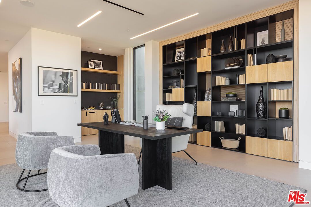 This sophisticated home office features a sleek, dark wood desk centered in the room, complemented by two modern grey upholstered armchairs. A large, floor-to-ceiling shelving unit with a mix of open black metal frames and light wood cabinetry provides ample storage and display space. The room is illuminated by minimalist recessed linear lighting, creating a clean, professional, and contemporary atmosphere.
