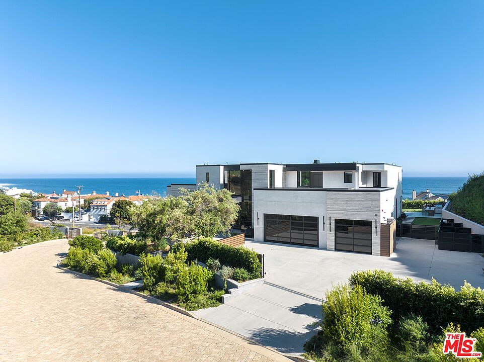 This modern, multi-level luxury residence features a clean, geometric architectural design with a light-colored stone facade and dark-framed windows. The property includes a spacious concrete driveway leading to a two-car garage, set against a stunning backdrop of the Pacific Ocean. The foreground is landscaped with lush greenery and a paved walkway, creating a sophisticated and serene coastal aesthetic.