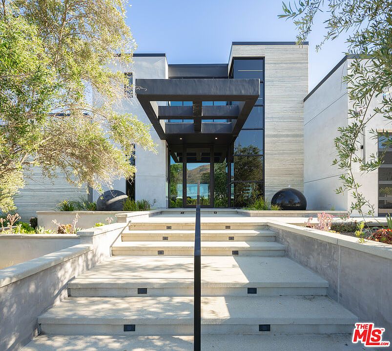 This striking modern residence features a grand, geometric entryway framed by a bold, dark architectural overhang and expansive glass panels. A set of wide, concrete steps with integrated lighting leads up to the front door, flanked by minimalist landscaping and spherical decorative accents. The home's facade combines clean white surfaces with textured stone, creating a sophisticated and contemporary aesthetic.