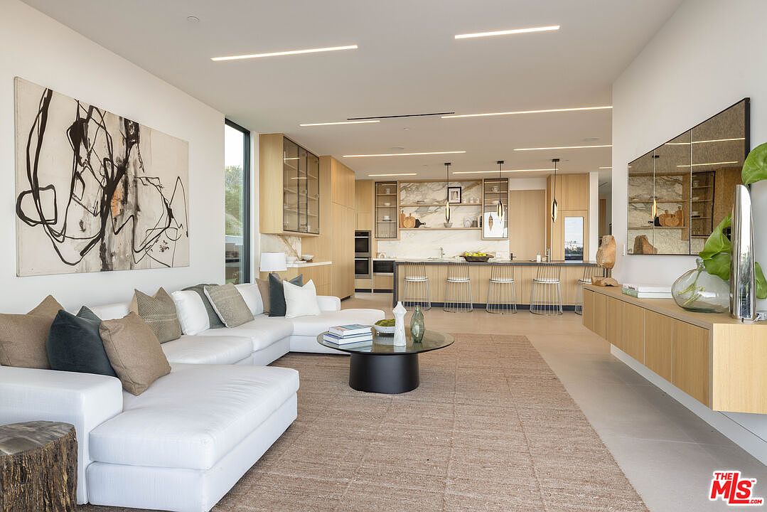 This expansive, open-concept living area showcases a modern aesthetic with a large white sectional sofa, a circular glass coffee table, and a striking abstract art piece on the wall. The space flows seamlessly into a sleek, light-wood kitchen featuring a long island with bar seating and recessed linear lighting throughout the ceiling. The perspective is captured from a low, eye-level angle, emphasizing the airy, sophisticated design and the harmonious blend of textures and neutral tones.