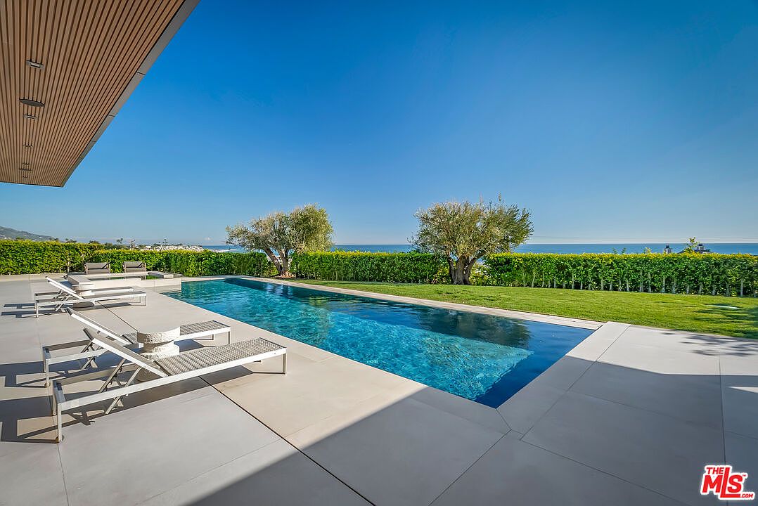 This stunning outdoor space features a sleek, rectangular infinity-edge swimming pool that seamlessly blends with the horizon of the ocean in the background. The expansive concrete patio is furnished with modern lounge chairs, providing a perfect vantage point for the manicured lawn and mature olive trees. The overall aesthetic is one of luxurious, contemporary coastal living, emphasizing clean lines and an open, airy atmosphere.