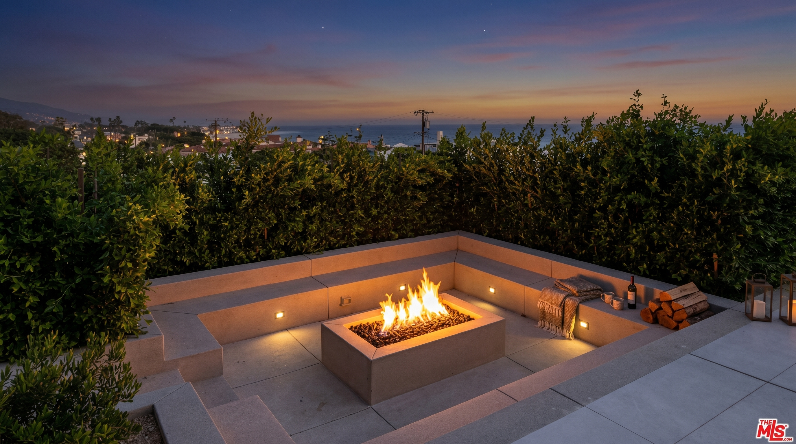 This outdoor space features a modern, built-in concrete fire pit surrounded by integrated bench seating, creating a sleek and minimalist lounge area. The patio is framed by lush, tall green hedges that provide privacy while framing a stunning, unobstructed panoramic view of the ocean and coastline. The perspective is eye-level, capturing the serene atmosphere of a high-end coastal retreat perfect for sunset gatherings.