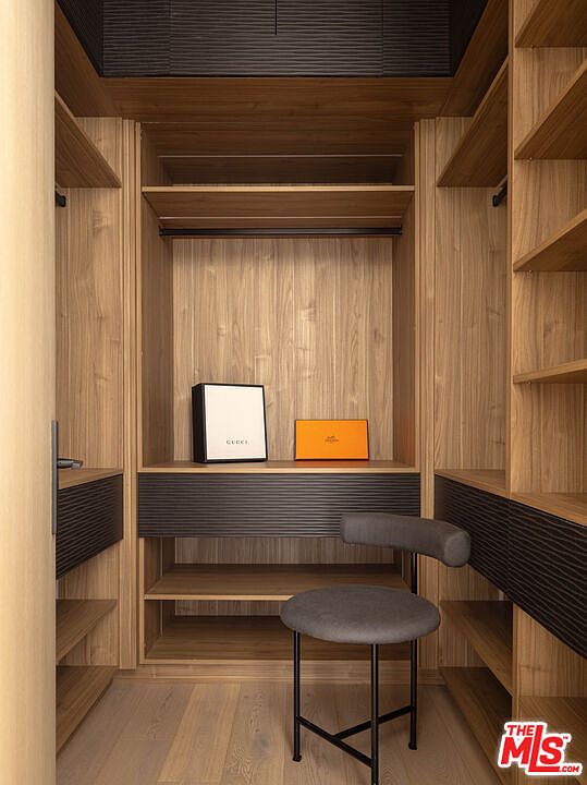 This sophisticated walk-in closet features custom light-wood cabinetry with integrated dark, textured drawer fronts and shelving. A central vanity area is accented by a modern, minimalist grey stool, while luxury brand boxes add a touch of elegance to the space. The perspective is a straight-on, symmetrical shot that highlights the clean lines and high-end craftsmanship of the custom storage design.