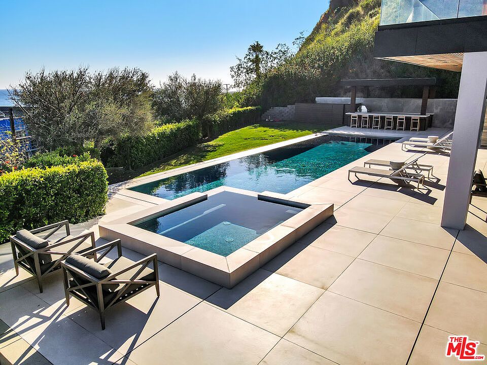 This luxurious outdoor living space features a sleek, rectangular swimming pool and an adjacent square spa, both set into a large, light-colored stone patio. The area is complemented by modern lounge chairs, a covered outdoor bar with seating, and a lush green lawn bordered by manicured hedges. The elevated perspective captures the serene atmosphere of this private backyard oasis, highlighting its clean lines and seamless integration with the natural hillside landscape.