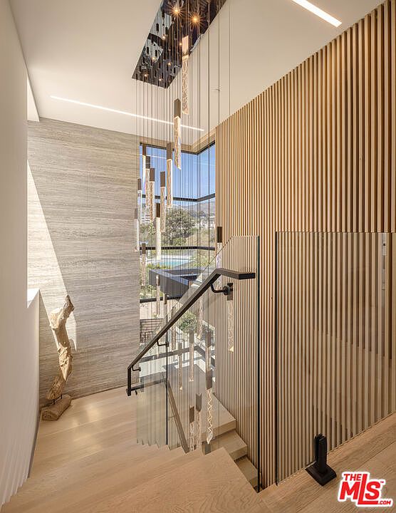 This sophisticated staircase area features a striking modern chandelier with cascading glass pendants that draw the eye downward. The space is defined by a dramatic contrast between a textured stone wall, a warm vertical wood-slat feature wall, and a sleek glass railing. The perspective from the top of the stairs highlights the open, airy design and the seamless integration of natural light from the large window.