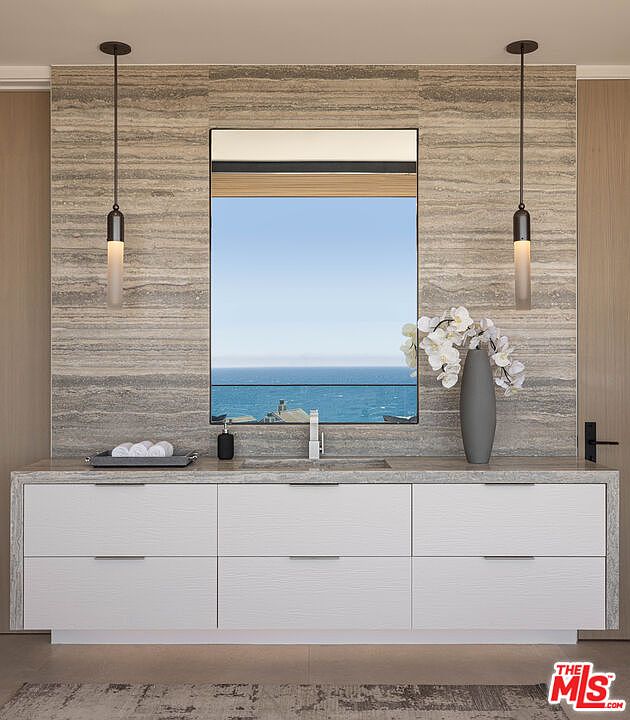 This luxurious primary bathroom vanity features a sleek, modern design with a textured stone backsplash and a large, frameless mirror reflecting a stunning ocean view. The white, multi-drawer vanity cabinet is topped with a matching stone countertop, complemented by minimalist pendant lighting and a decorative vase of white orchids. The overall aesthetic is sophisticated, serene, and high-end, emphasizing a seamless connection between indoor luxury and the natural coastal landscape.