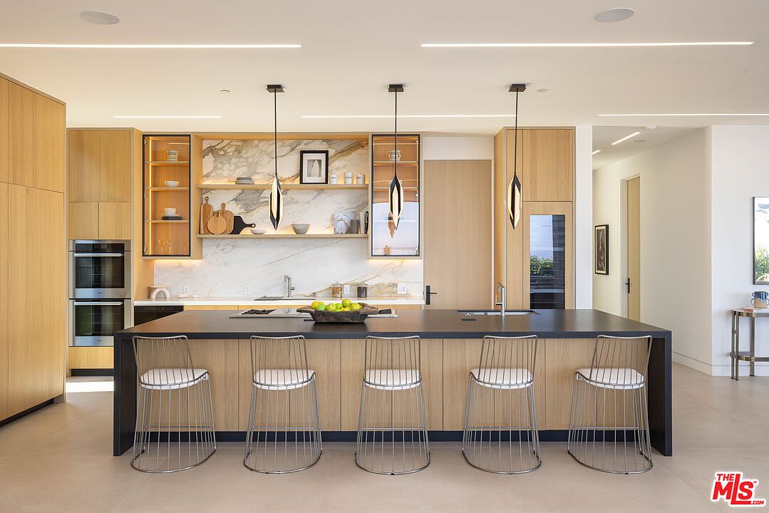 This modern, high-end kitchen features a large central island with a dark countertop and five sleek wire-frame bar stools. The space is defined by light wood cabinetry, a striking marble backsplash with open shelving, and minimalist pendant lighting. The overall aesthetic is clean, sophisticated, and contemporary, showcasing a seamless blend of natural textures and modern design.