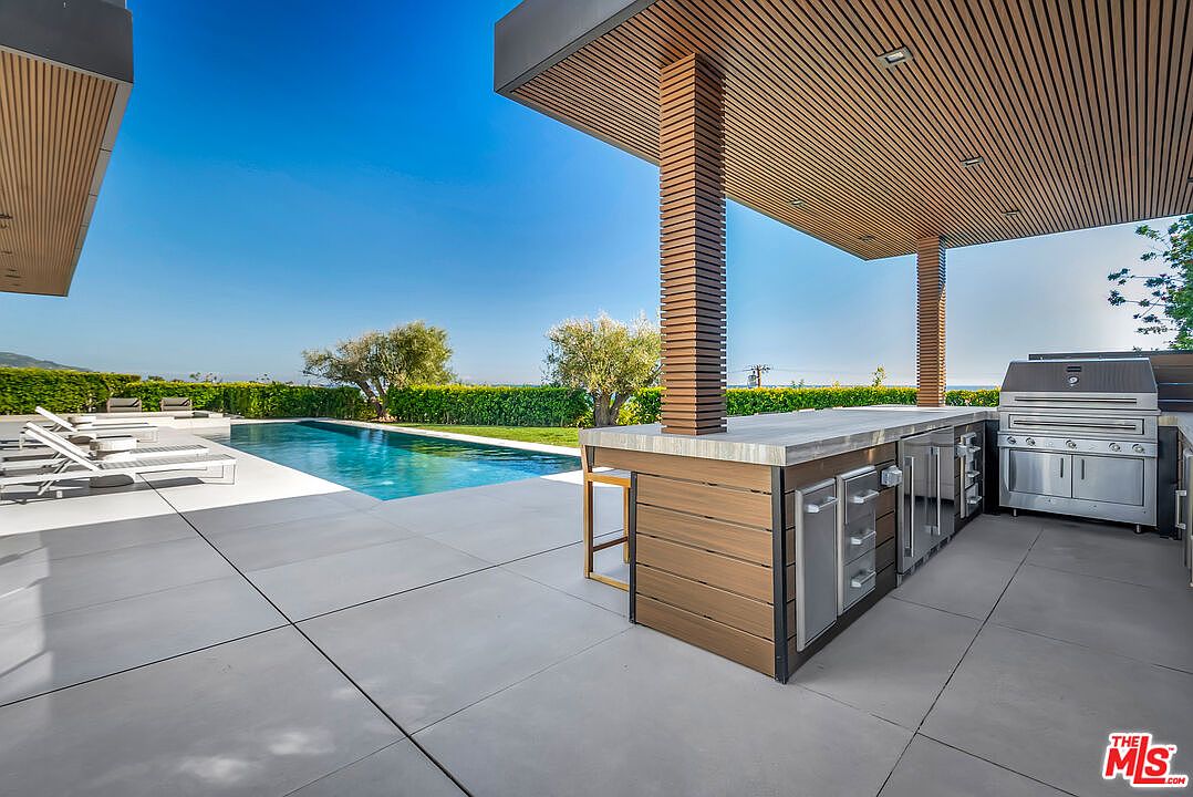 This luxurious outdoor living space features a sleek, modern infinity-edge pool set against a backdrop of lush green hedges and clear blue skies. A covered patio area includes a high-end stainless steel outdoor kitchen with a built-in grill and bar seating, perfect for entertaining. The clean lines of the concrete decking and the warm wood-paneled ceiling create a sophisticated, resort-style atmosphere.