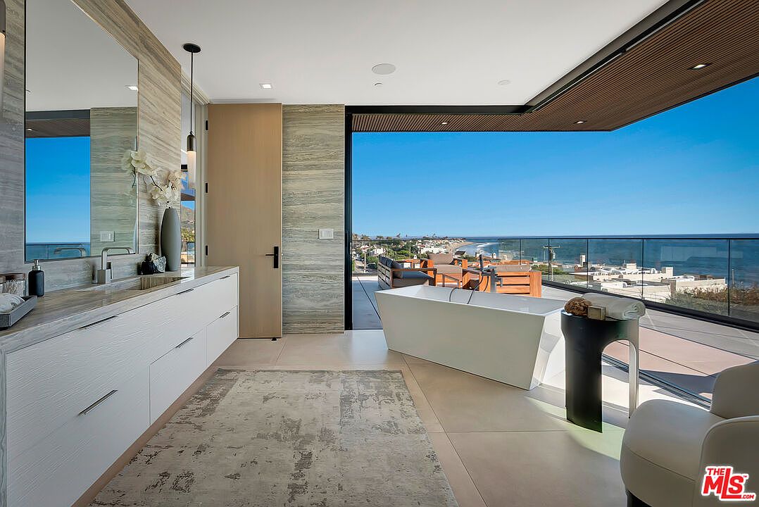 This luxurious primary bathroom features a sleek, modern design with a floating vanity, stone-textured walls, and a freestanding soaking tub positioned to overlook a stunning ocean view. The room seamlessly transitions to an outdoor balcony through floor-to-ceiling glass walls, creating an airy, open-concept atmosphere. The combination of high-end finishes and the panoramic coastal scenery provides a serene and sophisticated spa-like experience.
