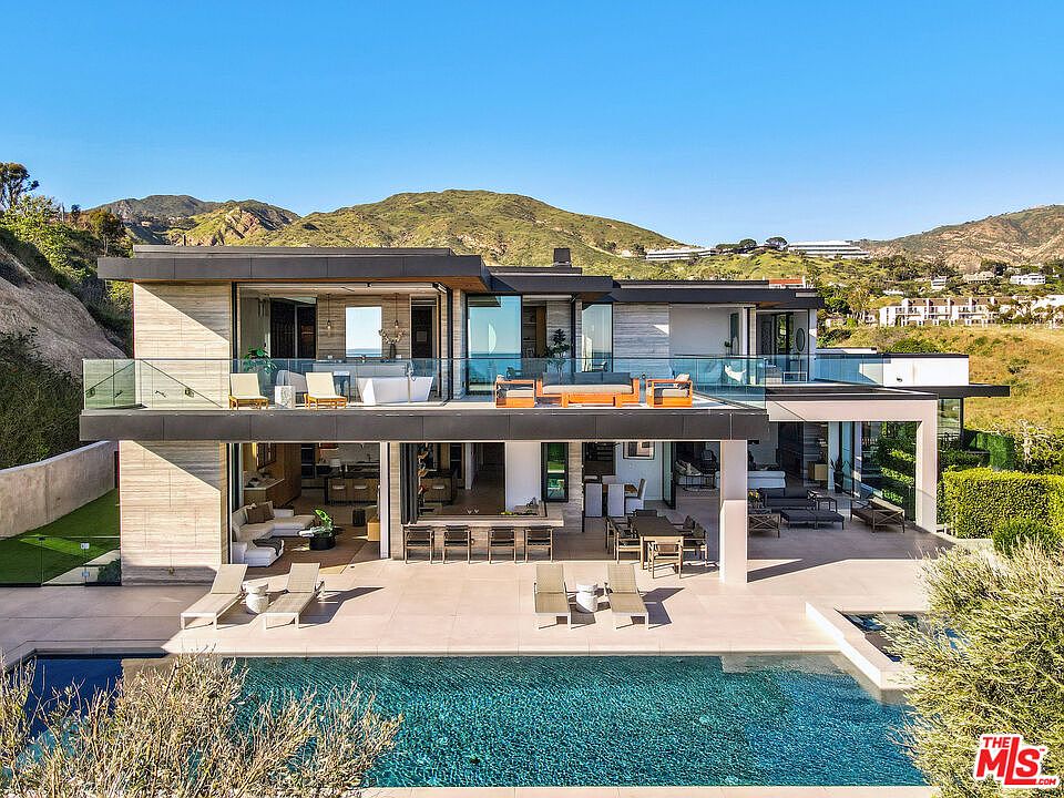 This stunning contemporary luxury residence features a seamless indoor-outdoor flow with expansive glass walls opening onto a multi-level terrace. The property boasts a sleek infinity-edge pool, a spacious patio area with outdoor dining and lounge furniture, and a second-story balcony overlooking the scenic hillside landscape. The modern architectural design emphasizes clean lines, natural stone textures, and an open-concept layout perfect for high-end entertaining.