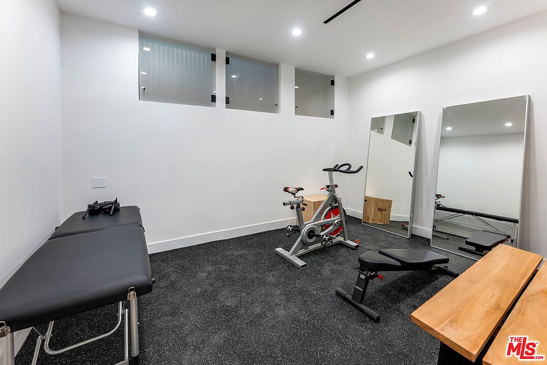 This modern home gym features a clean, minimalist aesthetic with white walls, recessed lighting, and dark, durable rubber flooring. The space is equipped with a stationary exercise bike, a weight bench, and a massage or treatment table, complemented by two large floor-to-ceiling mirrors that enhance the room's depth. High, frosted windows provide natural light while maintaining privacy, creating a bright and functional workout environment.