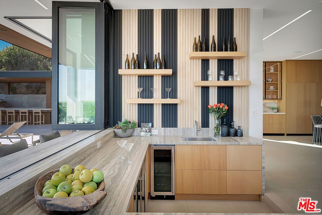 This sophisticated wet bar area features a seamless blend of natural wood cabinetry and a striking vertical slat wall with contrasting black and light wood tones. The space is equipped with a built-in wine refrigerator, a sleek sink, and open shelving displaying glassware and bottles, all set against a beautiful stone countertop. The perspective captures an inviting indoor-outdoor flow, with large glass doors opening to a sun-drenched patio, creating a luxurious and functional entertainment space.