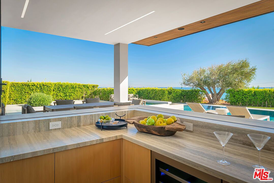 This image captures a sleek, modern outdoor kitchen area that seamlessly blends indoor luxury with an open-air environment. The space features high-end stone countertops, a wooden bowl filled with fresh fruit, and a wine refrigerator, all set against a stunning backdrop of a swimming pool and a lush green hedge. The perspective is from behind the bar, looking out toward the sunny patio and ocean horizon, creating an inviting and sophisticated atmosphere for entertaining.