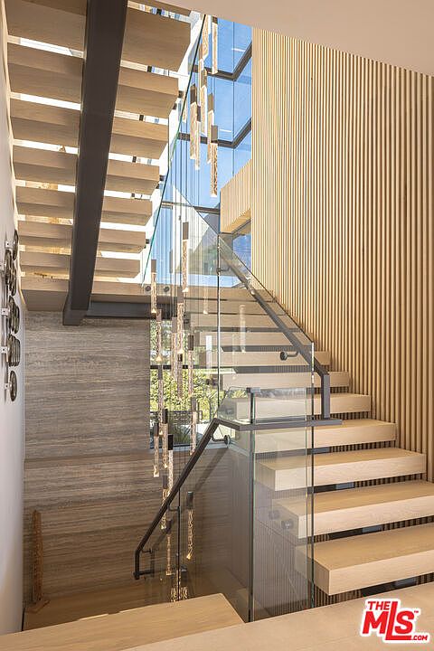 This striking interior staircase features floating light-wood treads and a sleek glass railing, creating an open and airy architectural statement. A dramatic, multi-tiered crystal chandelier hangs vertically through the stairwell, complemented by a textured wood-slat accent wall and large floor-to-ceiling windows that flood the space with natural light. The perspective is captured from a slightly elevated angle, emphasizing the modern, luxurious design and the seamless integration of natural materials.